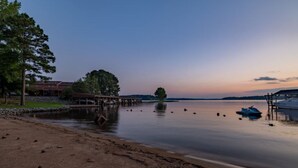 Vista del lago con un muelle de madera que se extiende hacia el agua, un barco amarrado a la derecha y un edificio con un árbol grande al fondo.