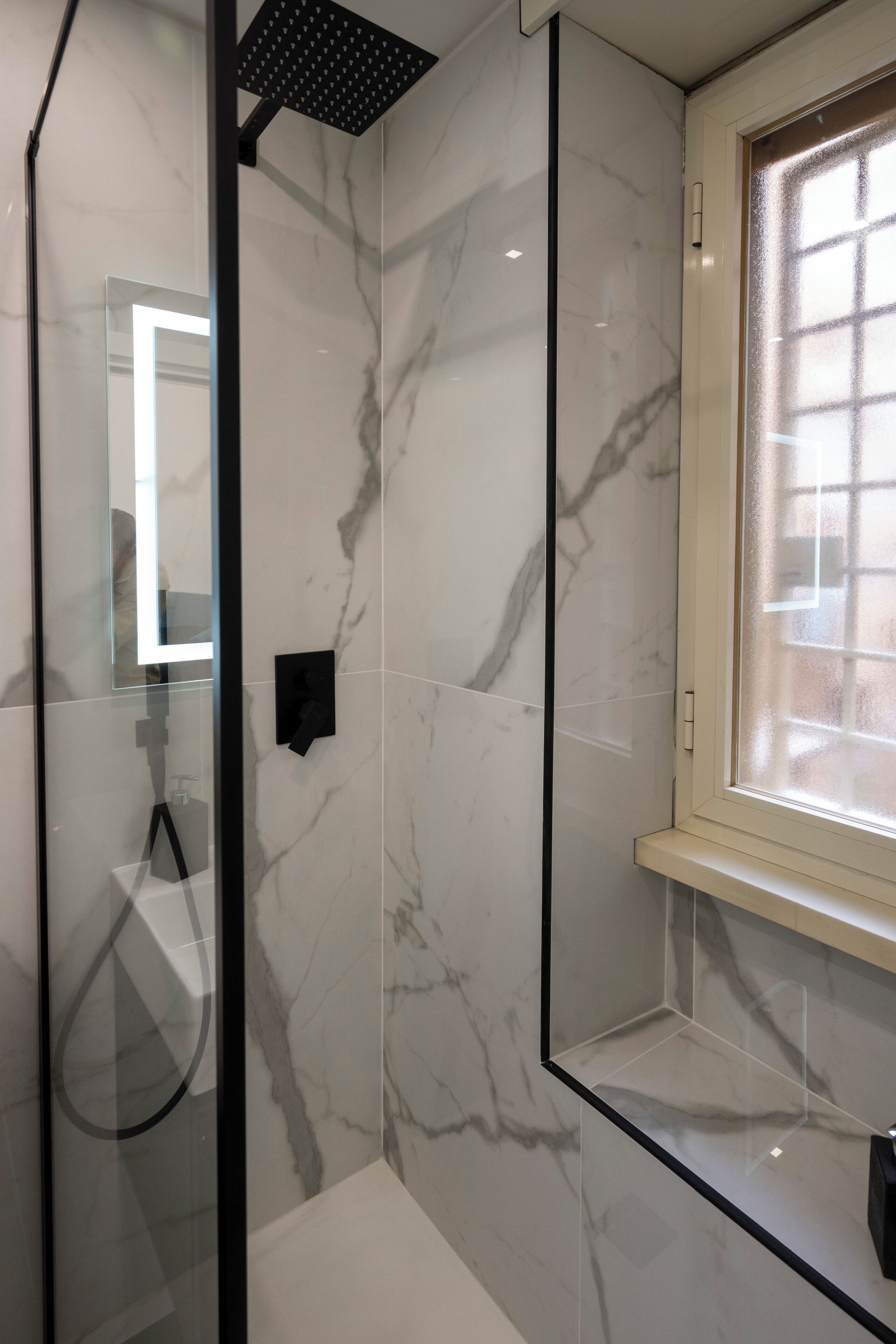 A modern bathroom with a glass-enclosed shower, marble walls, and a window.