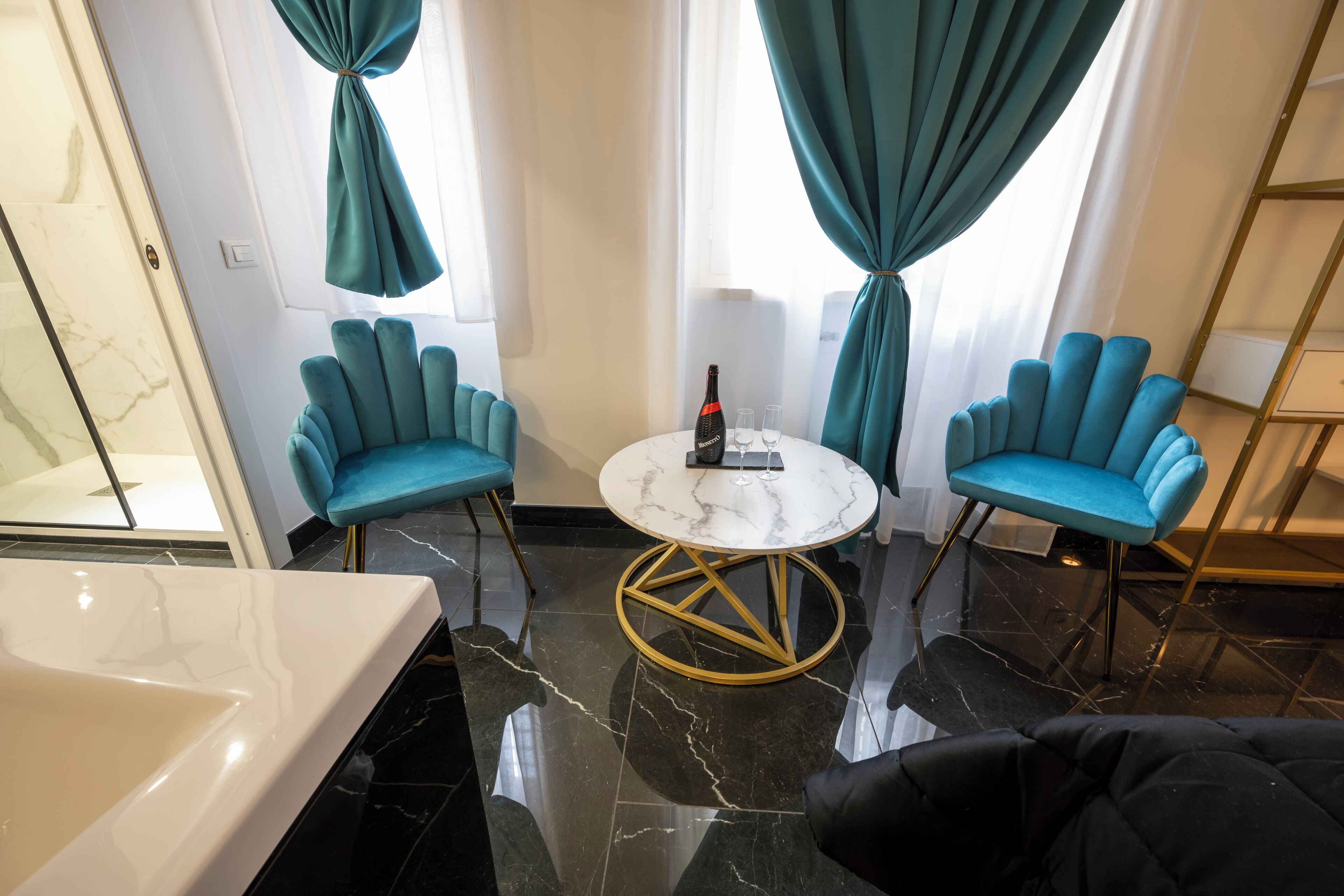 Une chambre d’hôtel moderne avec un sol en marbre, une baignoire blanche, deux fauteuils turquoise, une table ronde avec une bouteille et des verres, et une grande fenêtre ornée de rideaux turquoise.