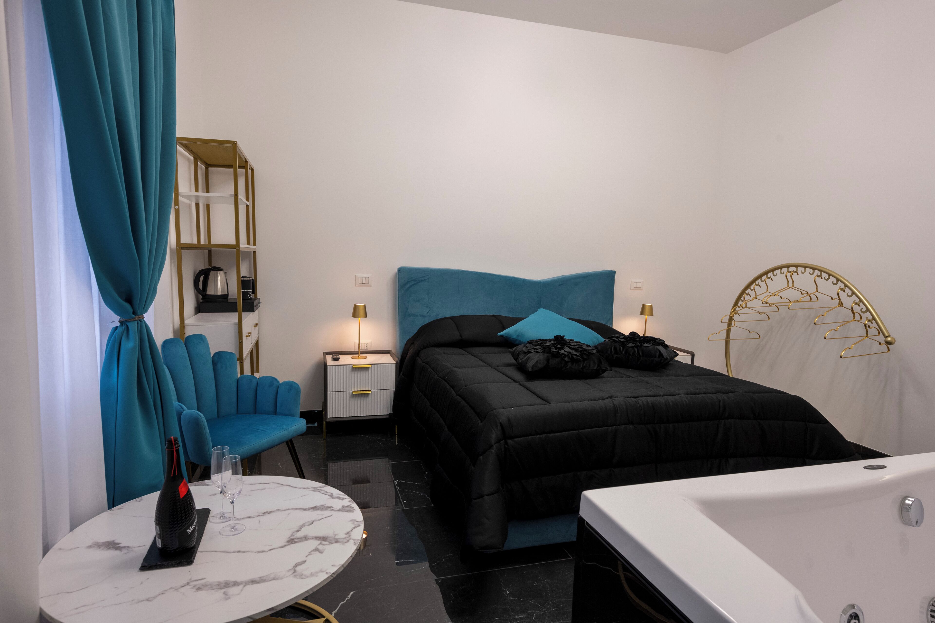 A modern bedroom with a black bed, teal curtains, a white marble table with a wine bottle, and a white bathtub.