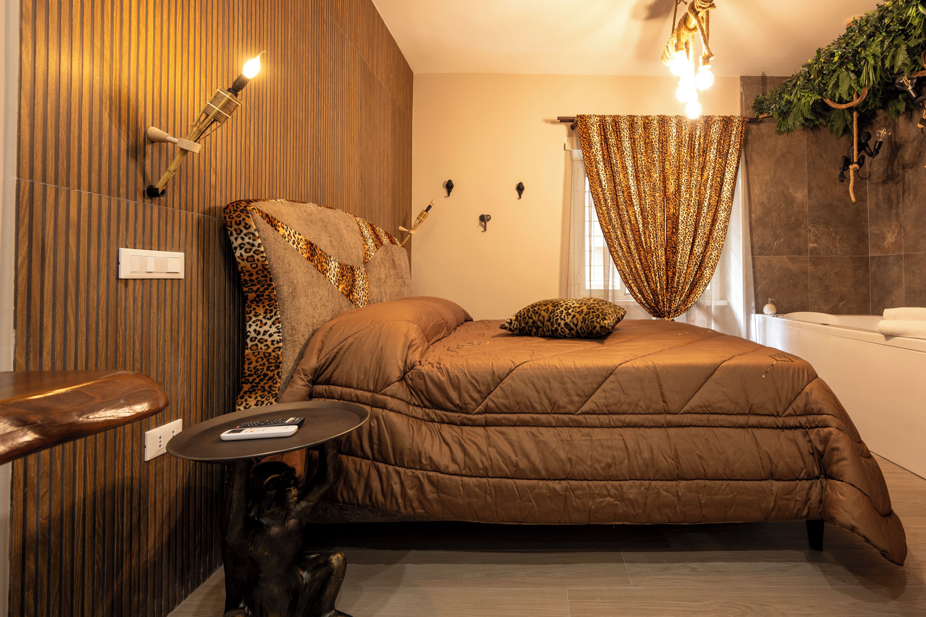 Une chambre avec un lit marron, une petite table ronde avec une télécommande, un luminaire fixé au mur et un rideau orné d’un motif de feuilles.