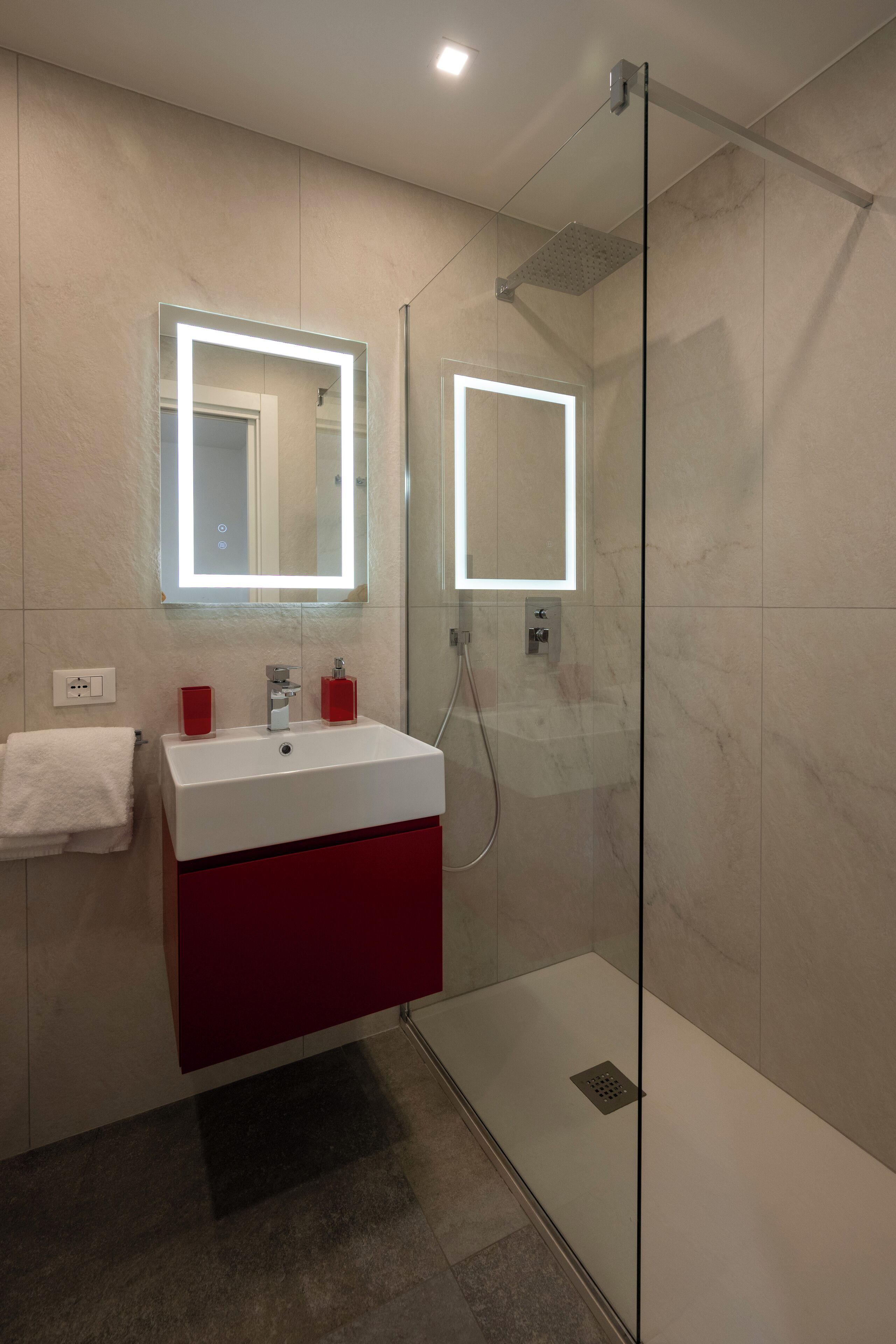 A modern bathroom with a glass-enclosed shower, a white sink with red accents, and a large mirror with integrated lighting.
