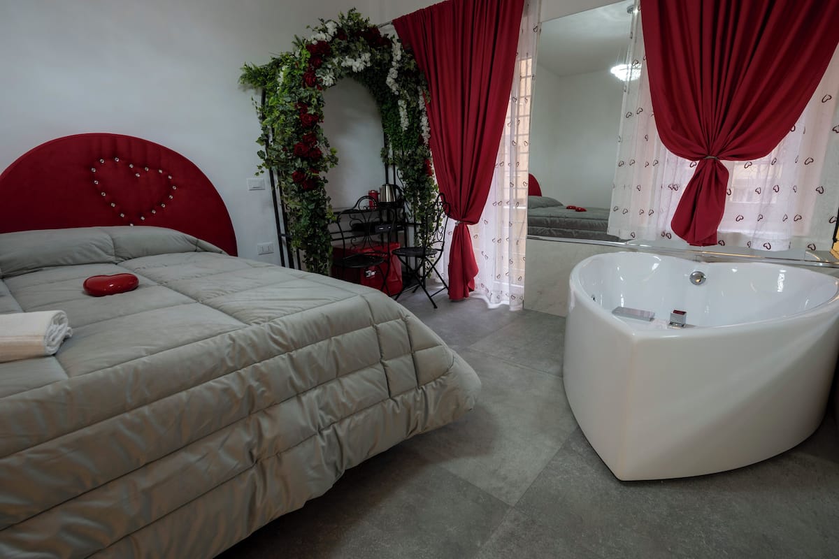 A bedroom with a large bed, a red heart-shaped headboard, a white bathtub, and a shower area.