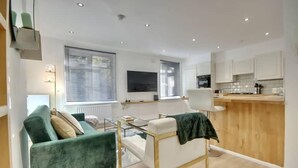 Ein modernes Wohnzimmer mit einem grünen Sofa, einem weißen Sessel und einem Glastisch. Es gibt eine Küchenzeile mit weißen Schränken und einer Holz Arbeitsplatte.