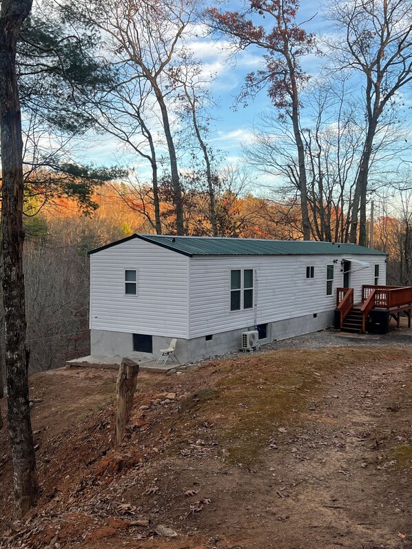 A single-story mobile home with a green roof, a small porch with a wooden bench, and a dirt yard.