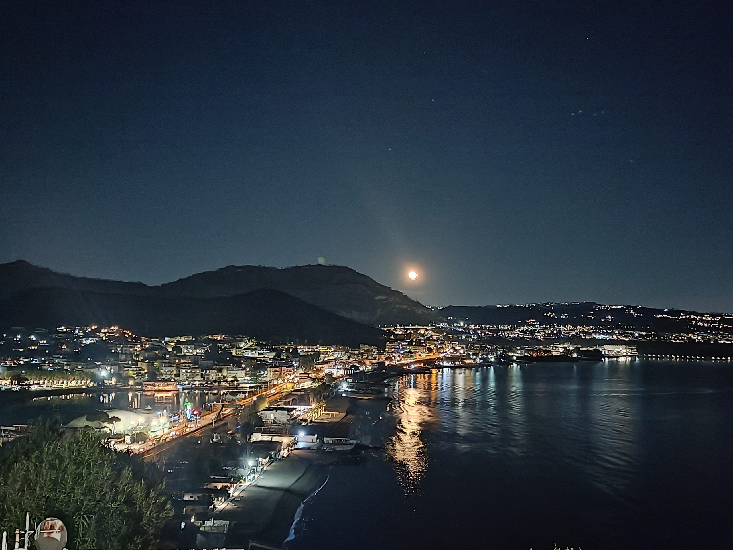 Un pueblo costero de noche con luna visible, agua tranquila que refleja luces y una montaña al fondo.