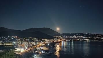 Un pueblo costero de noche con luna visible, agua tranquila que refleja luces y una montaña al fondo.