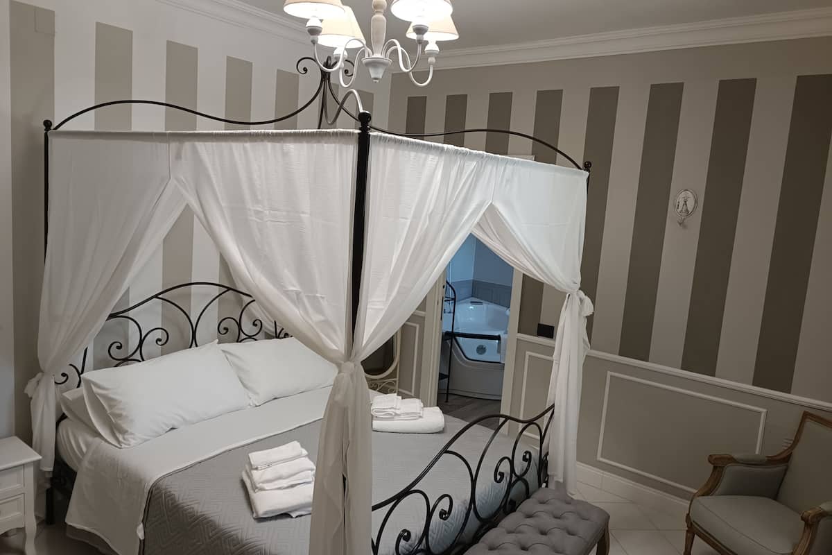 A four-poster canopy bed with white linens and a matching bench, a bedside table, a chair, and a chandelier.