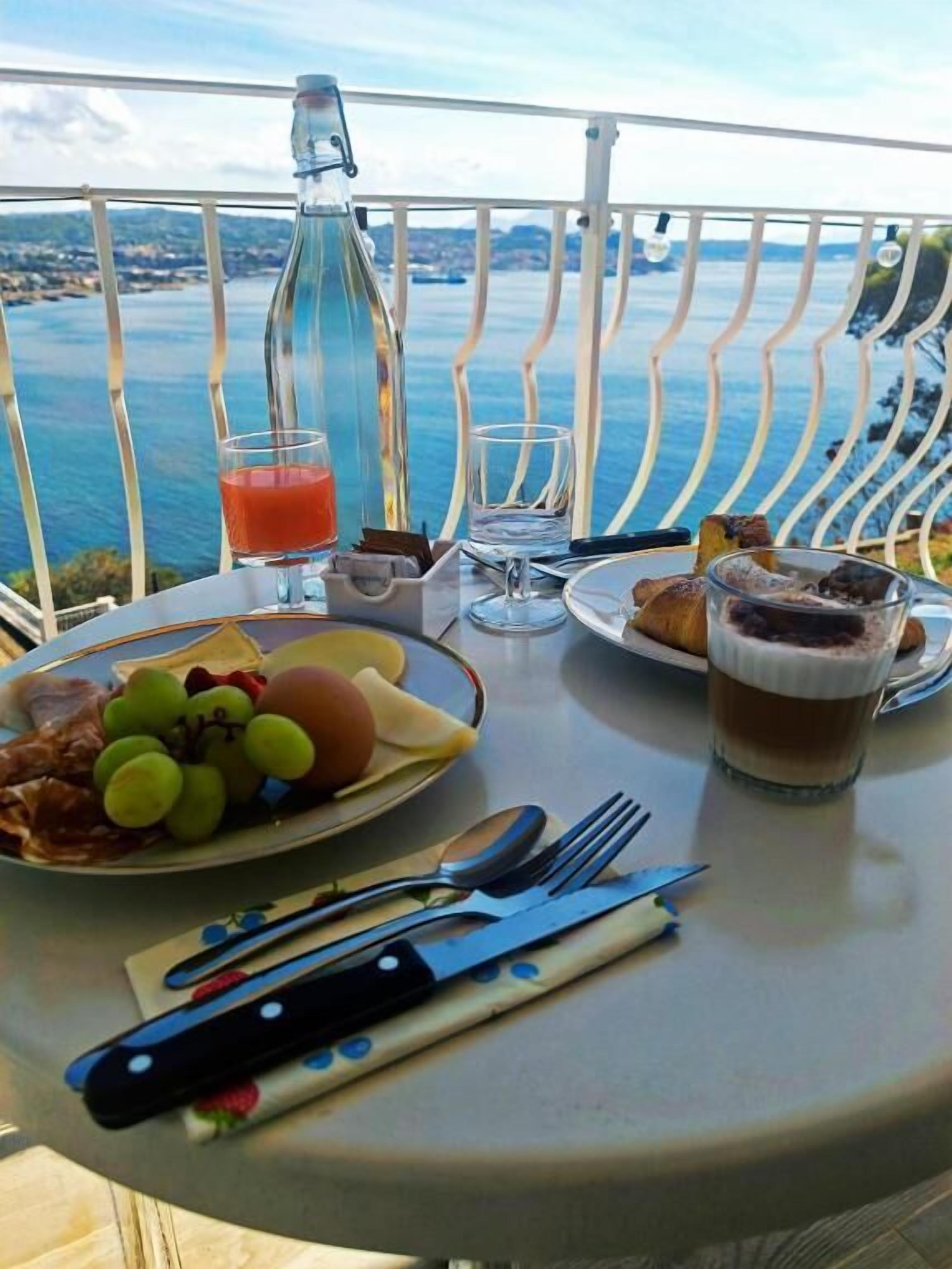 A table with breakfast items, a bottle, and a glass of orange juice on a balcony with a view of the sea.