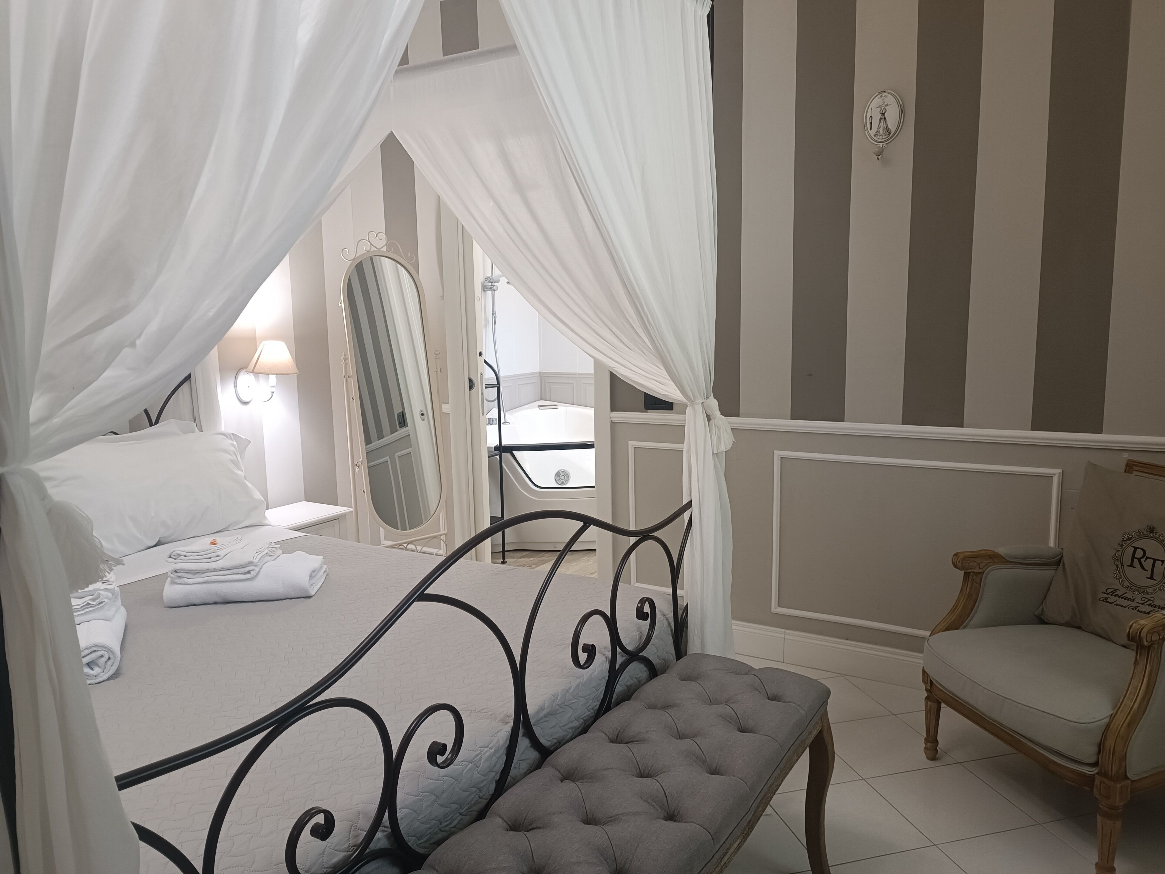 A bedroom with a four-poster bed, a bedside table with a lamp, a chair, and a large mirror.