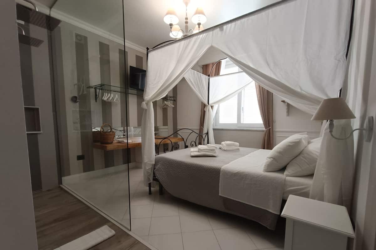 A bedroom with a canopy bed, a bedside table, a mirror, and a window with curtains.