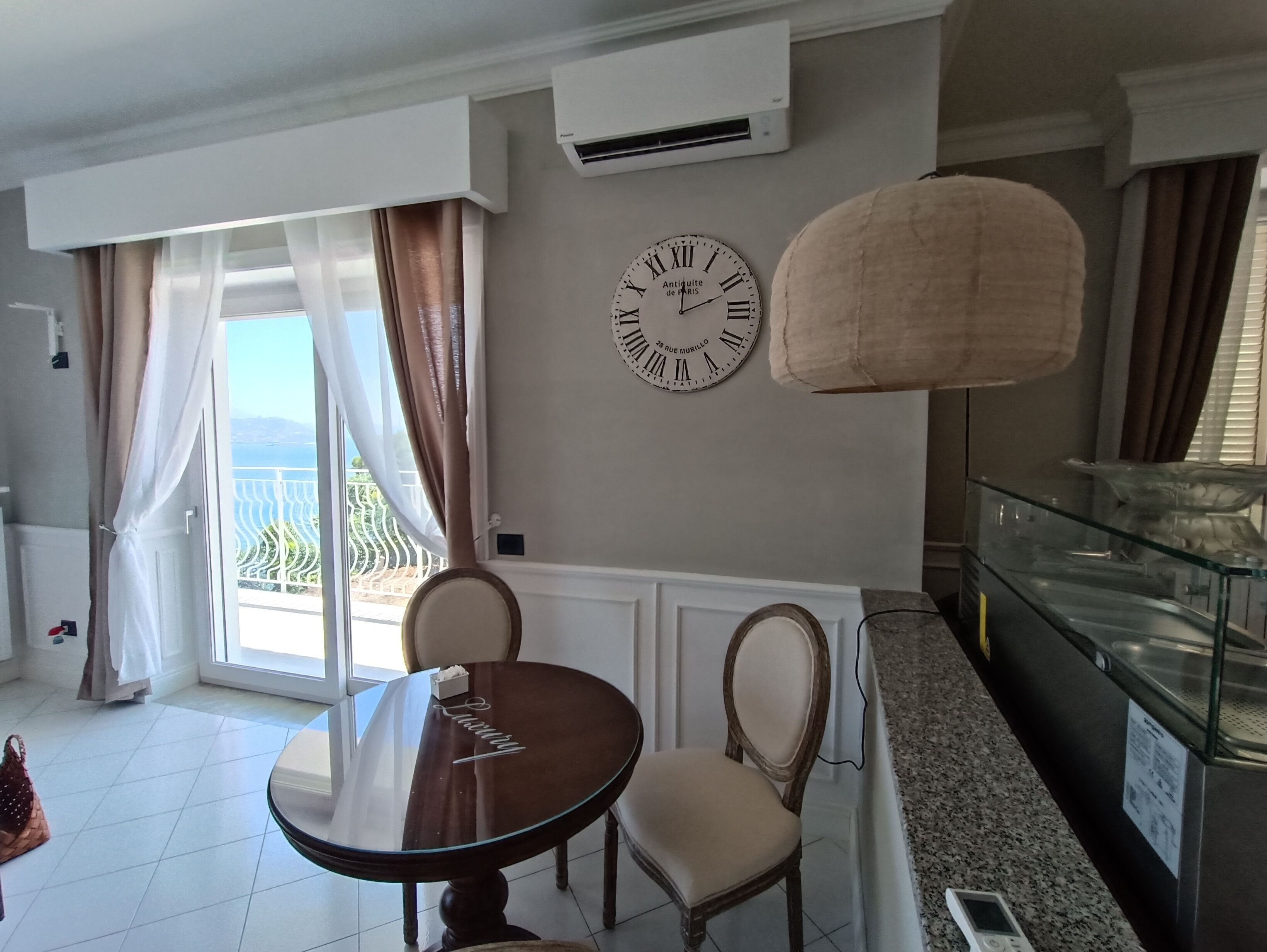 A room with a dining table, two chairs, a wall clock, an air conditioning unit, and a view of the outdoors.