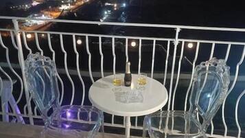 Un balcón con una mesa blanca y dos sillas transparentes, con vistas a un paisaje urbano nocturno.