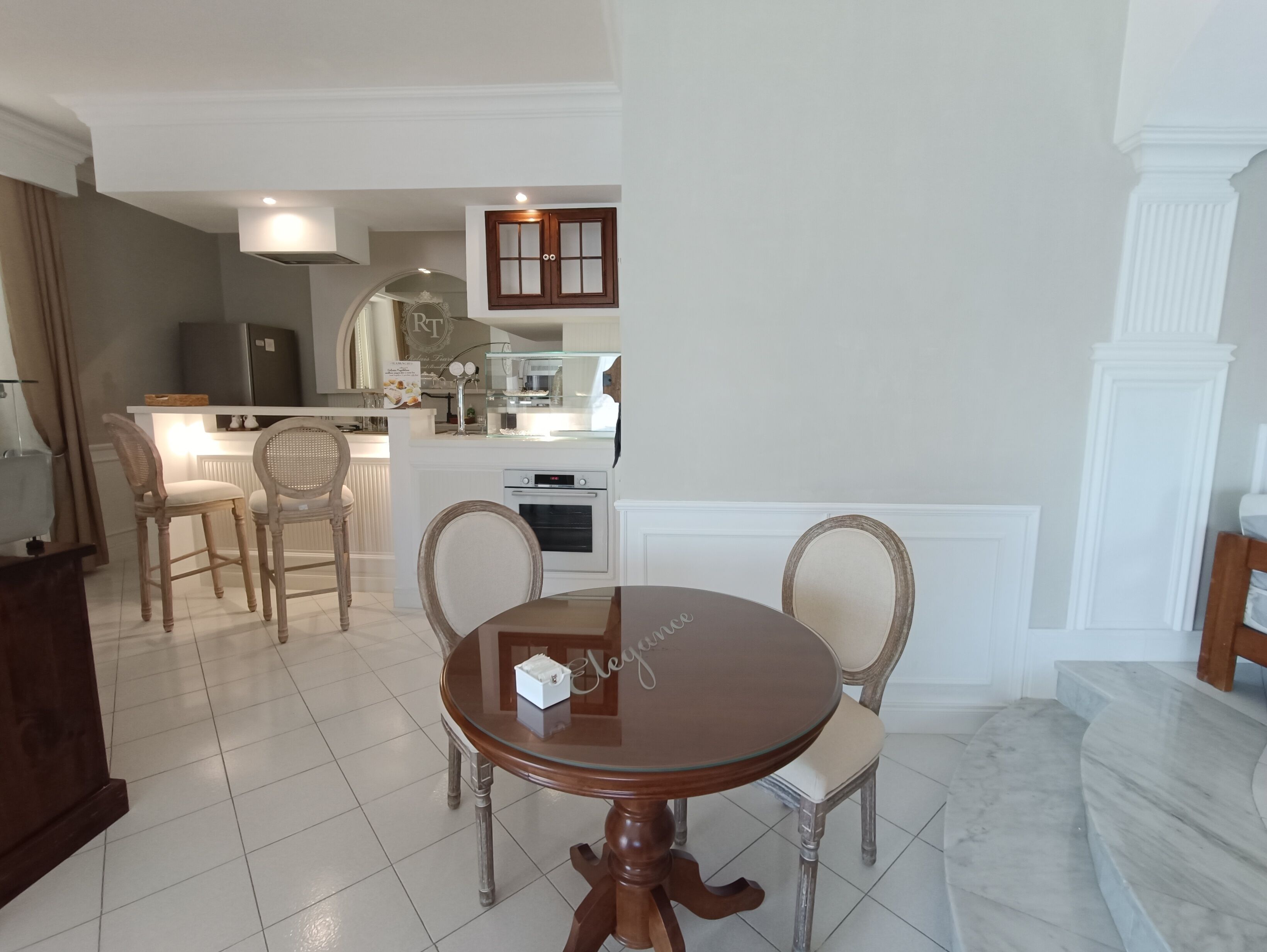 A dining area with a round table and four chairs, a kitchen in the background, and a white column.