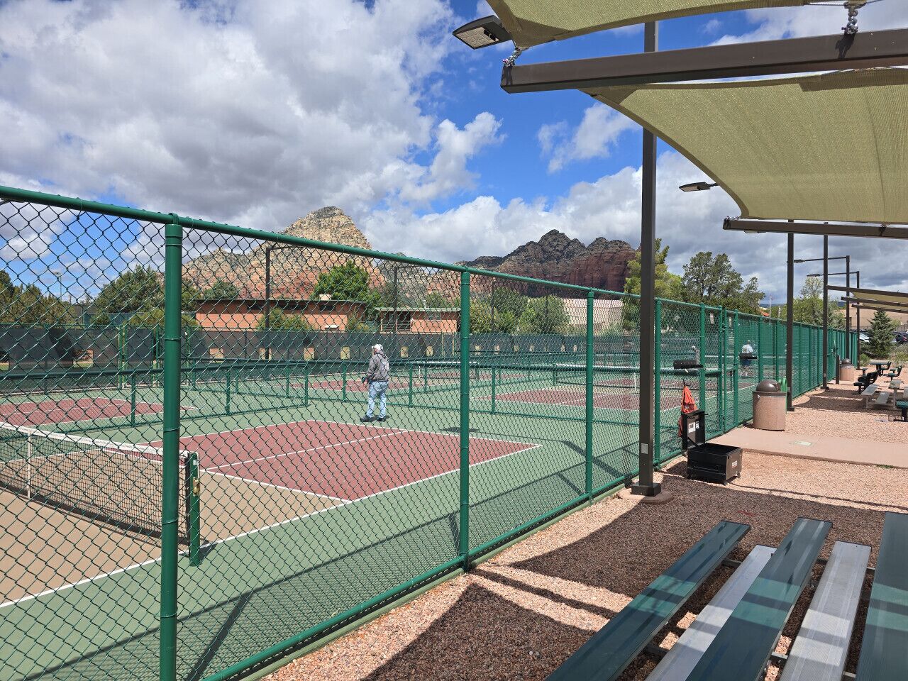 A tennis court enclosed by a green fence, with a bench and trash can nearby.