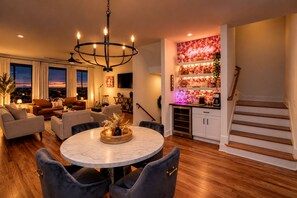 A modern living room with a round marble table, dark upholstered chairs, a chandelier, and a city view through large windows.