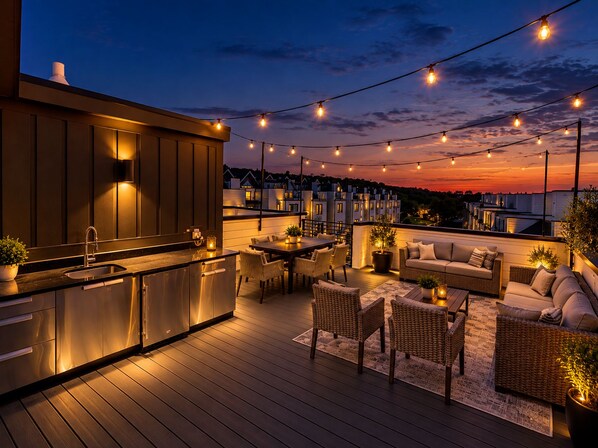 A rooftop deck with a bar area, outdoor seating, and string lights against a sunset backdrop.