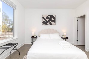 Ein modernes Schlafzimmer mit einem großen Bett, zwei Nachttischen mit Lampen, einem Fenster mit Blick auf Bäume und einer Tür, die in einen anderen Raum führt.