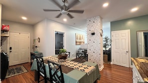 Un coin repas avec une table dressée pour quatre personnes, un ventilateur de plafond et un espace cuisine équipé d’un réfrigérateur et de placards.