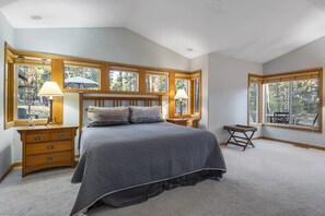 Un dormitorio con una cama grande, cabecera de madera, dos mesitas de noche con lámparas y vistas al exterior a través de amplias ventanas.