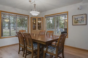 A dining room with a large wooden table, six chairs, a chandelier, and large windows with a view of trees.