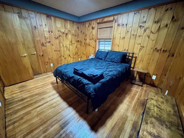 A bedroom with wooden panel walls, a bed with a dark blue bedspread, and a window with blinds.