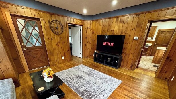 Un salon chaleureux avec des murs en bois, un grand téléviseur à écran plat, un canapé gris et une table basse ornée d’un vase de fleurs.