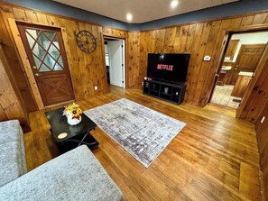 A cozy living room with wooden walls, a large flat-screen TV, a grey sofa, and a coffee table with a vase of flowers.
