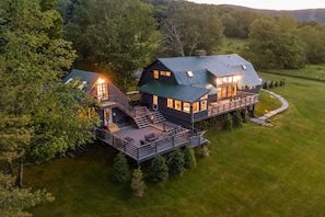 A large, two-story house with a deck and multiple windows, surrounded by trees and a grassy field.