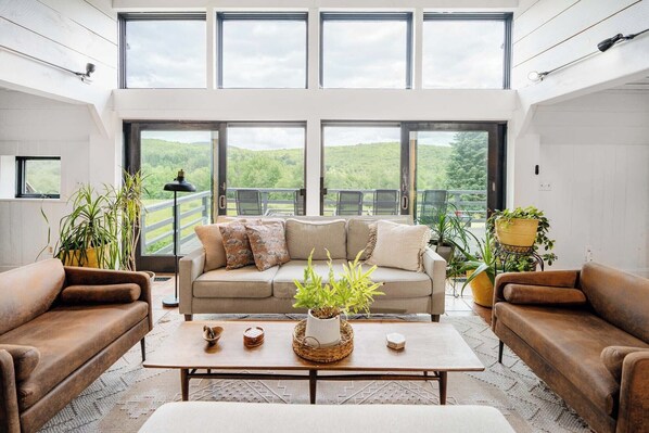 Een ruime woonkamer met een groot raam dat uitkijkt op een prachtig landschap, een beige bank, twee bruine fauteuils en een salontafel met decoratieve objecten.
