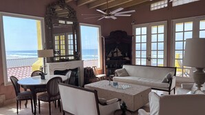 Ein sonnendurchflutetes Zimmer mit Meerblick, ausgestattet mit einer Couch, Sesseln, einem Tisch und einem Bücherregal.