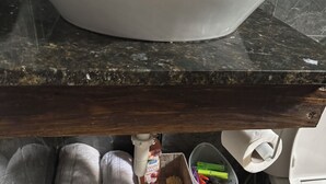 Un baño con encimera de mármol, un lavabo blanco, un estante de madera con toallas, una cesta y una bolsa de basura.