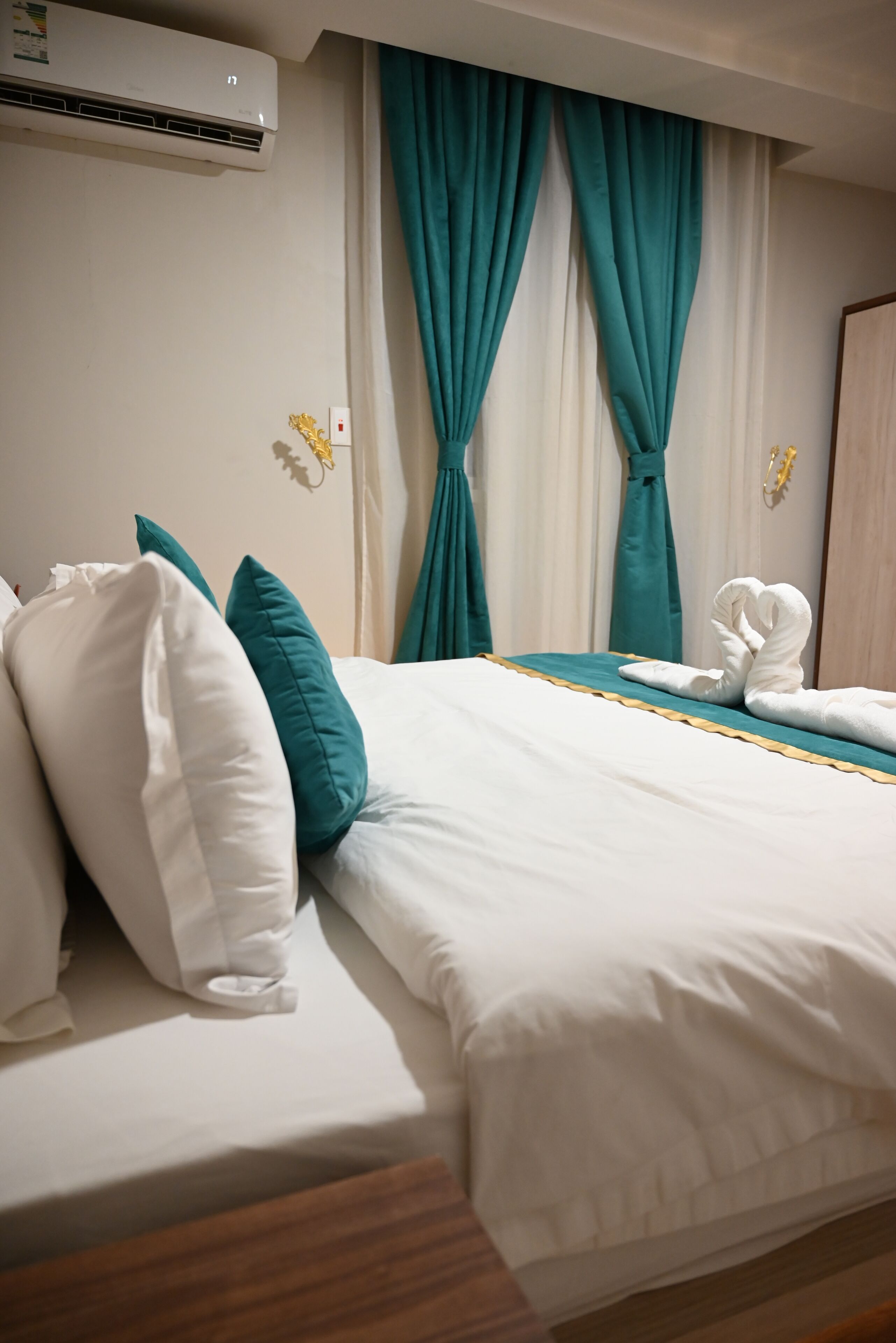 Una habitación de hotel con una cama, cortinas verde azulado, ropa de cama blanca y una toalla sobre la cama.