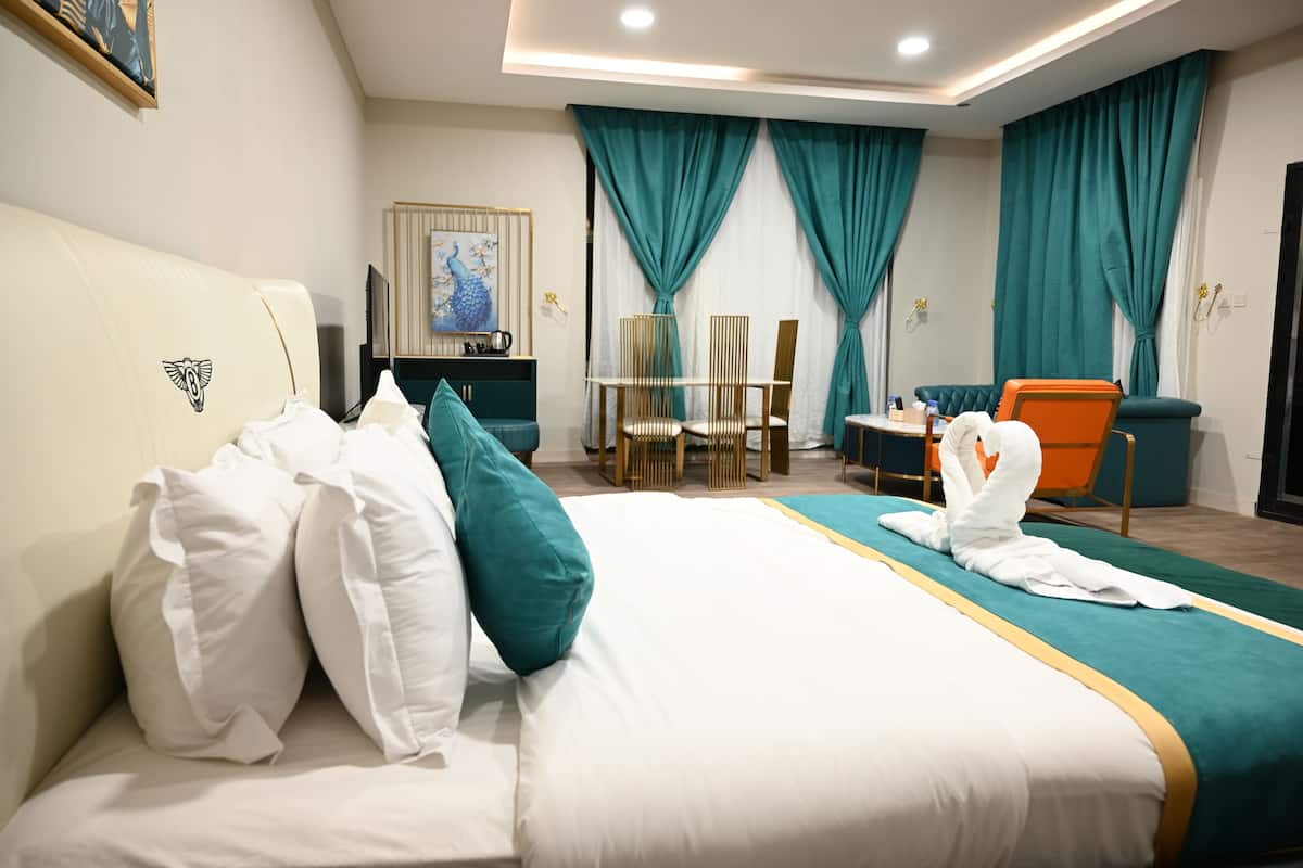 A hotel room with a large bed, teal curtains, a dining area with chairs, and a TV on a stand.