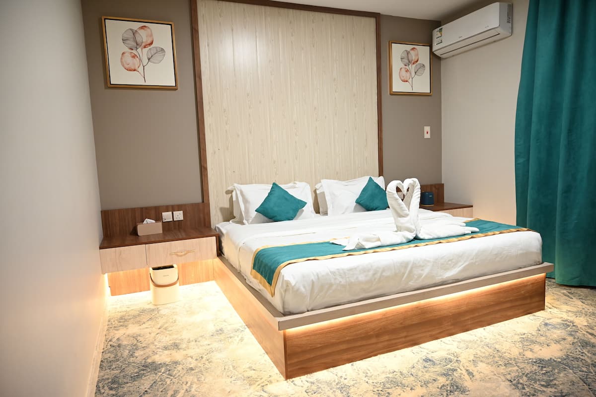 A neatly arranged hotel room with a large bed, a bedside table, a nightstand, a wall-mounted air conditioner, and a framed floral artwork on the wall.