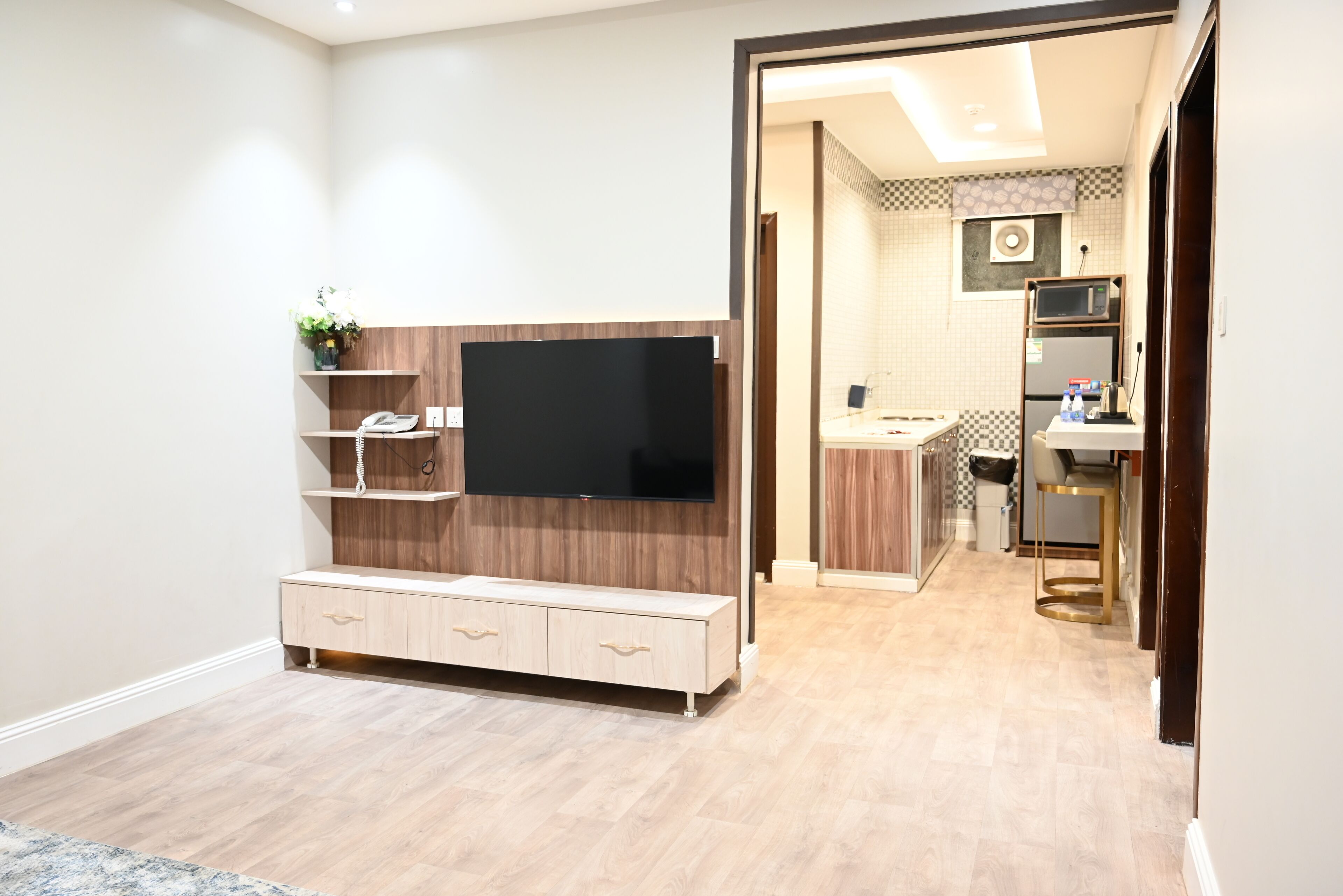 A modern living room with a flat-screen TV, wooden entertainment unit, and a kitchen area visible through an open doorway.