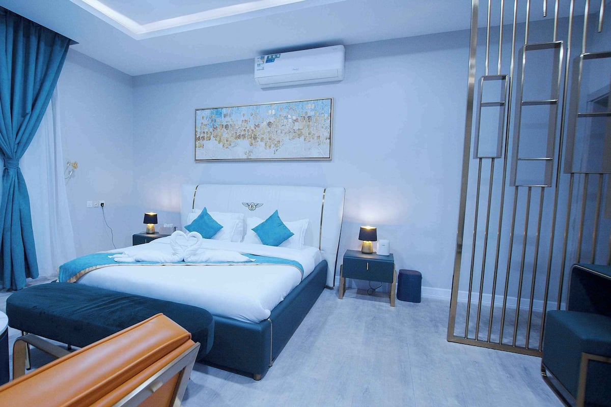 A modern hotel room with a large bed, a sitting area with a sofa and a table, a small bedside table with a lamp, and a wall-mounted air conditioner.