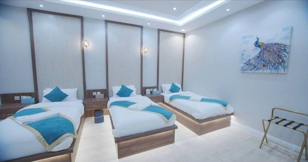 A hotel room with four single beds, each with a wooden headboard and white bedding with blue accents. There is a painting of a peacock on the wall and a small rack for hanging items.
