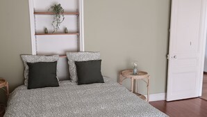 Un dormitorio con una colcha estampada, dos almohadas negras, una planta pequeña en un estante y dos mesitas de noche de mimbre con lámparas.