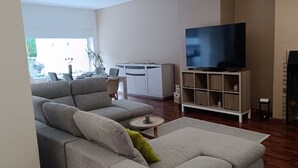 Una sala de estar con un sofá gris, un televisor sobre un mueble bajo, una chimenea y un comedor con mesa y sillas.