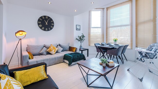 A modern living room with a sofa, coffee table, dining table, and chairs. A baby rocker is visible in the corner.