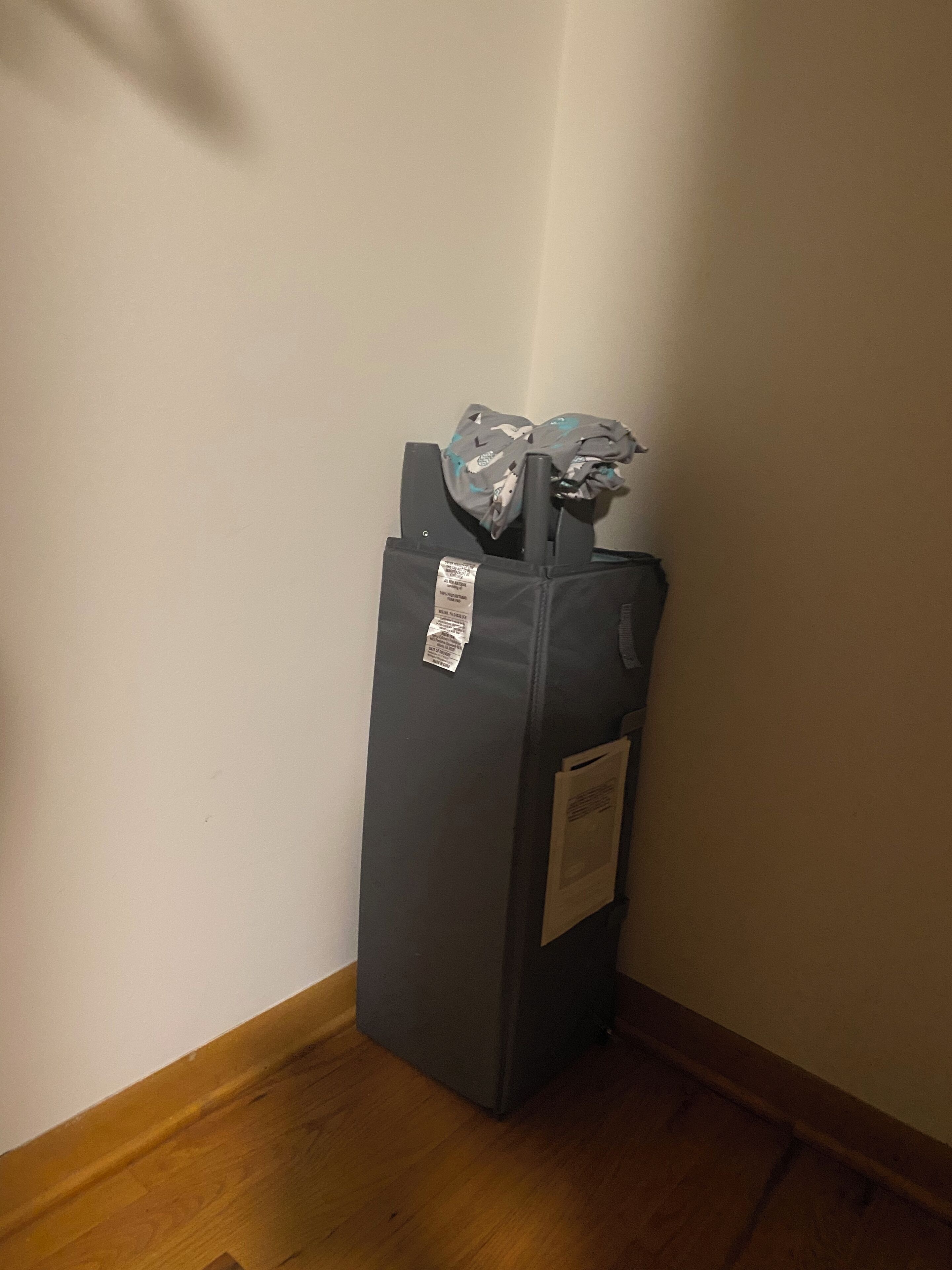 A corner of a room with a dark-colored trash bin against a white wall and wooden floor.