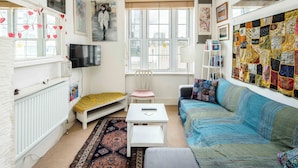 A cozy living room with a sofa, a small table, a chair, a window with heart decorations, a quilted wall hanging, and a television.