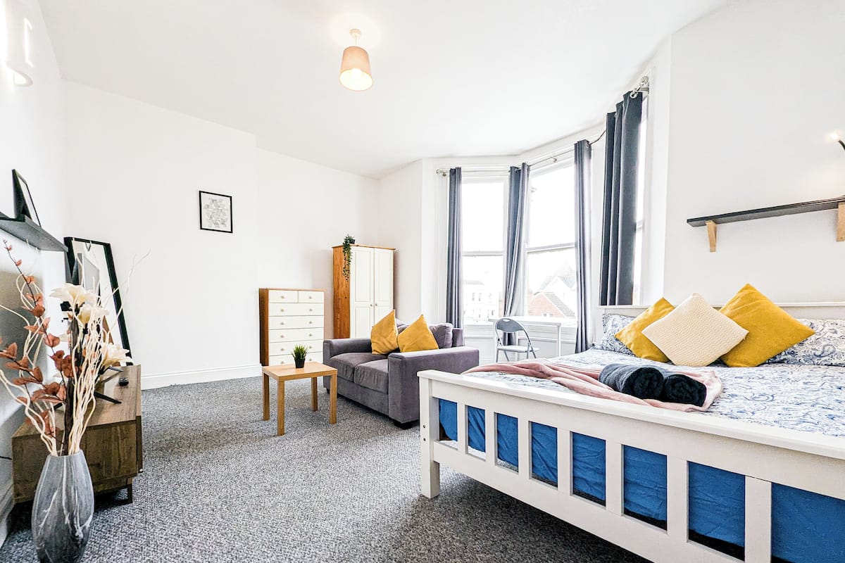 Un dormitorio con cama, sofá, una mesita, un armario y ventana con cortinas.