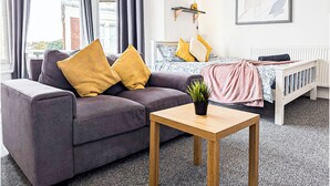 Een moderne woonkamer met een grijze bank, een houten salontafel, een bed met een wit hoofdbord en een lijst met een abstract schilderij aan de muur.