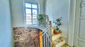 Una escalera con un muro de piedra, barandilla metálica, una planta en maceta y una puerta con un cartel.