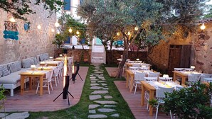 Un área de comedor al aire libre con muros de piedra, mesas y sillas de madera, un sendero de piedra y luces colgantes.