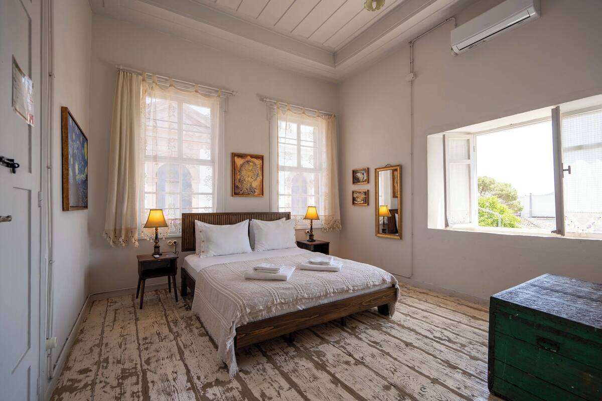 Quarto com cama de madeira, duas mesas de cabeceira com luminárias, cômoda verde e janela com cortinas.