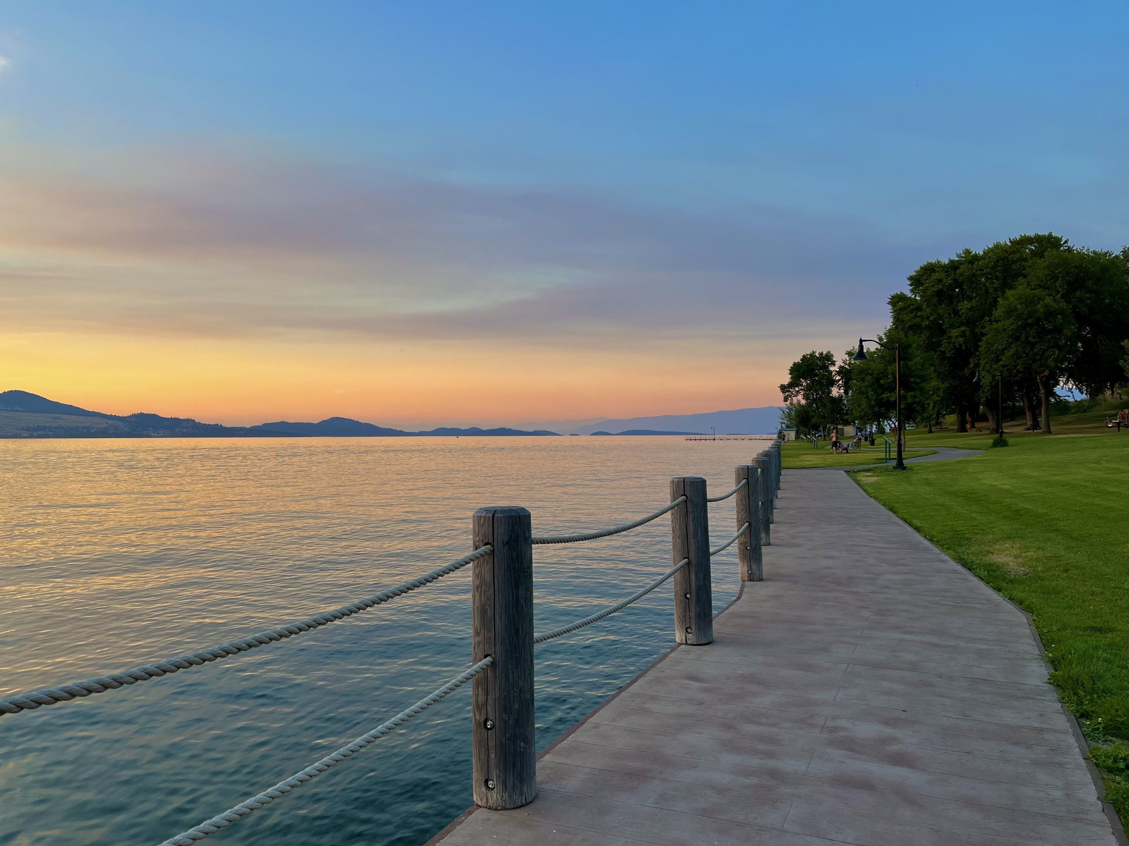 A waterfront walkway with a rope barrier, grassy areas, and trees.