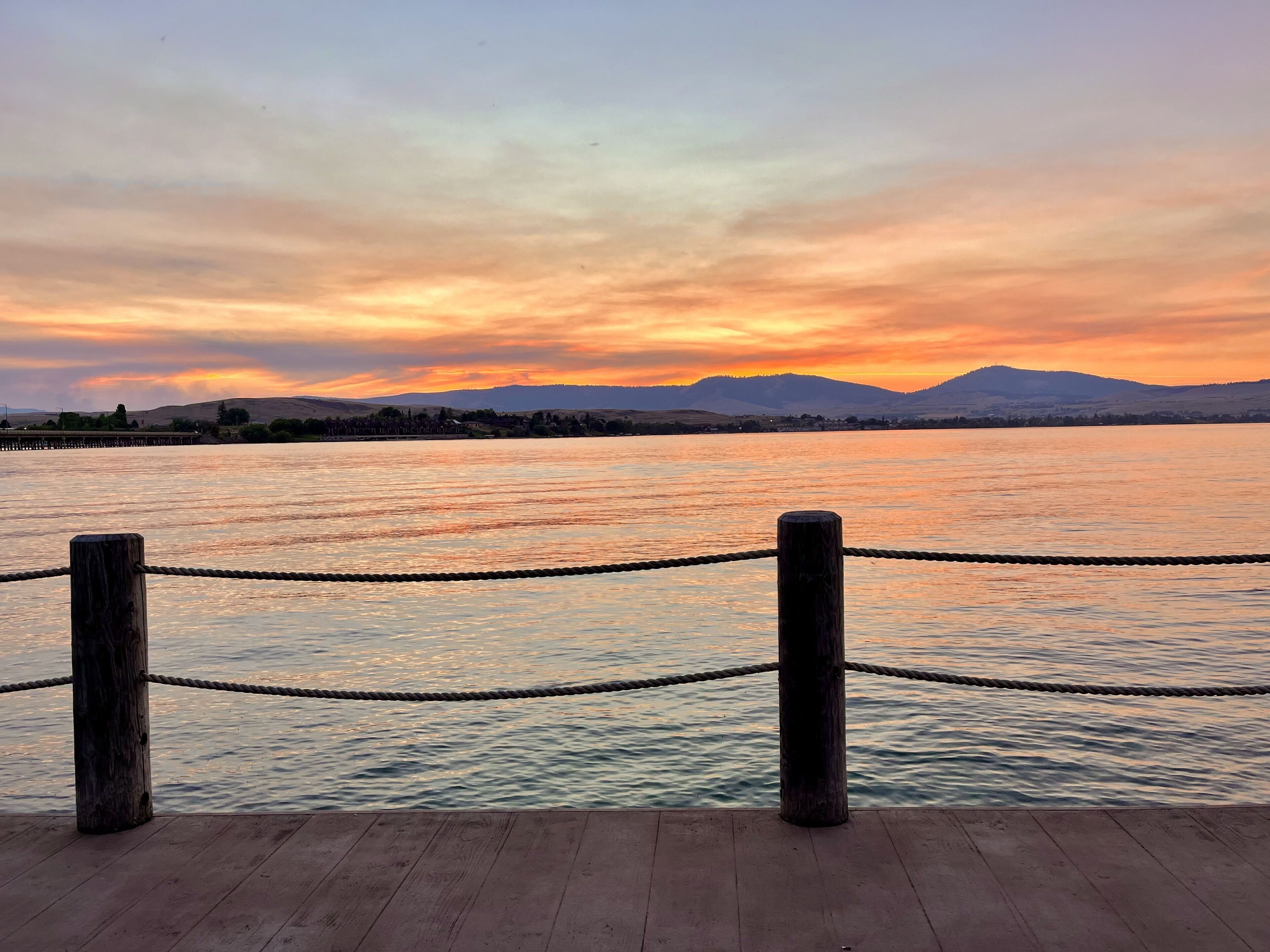 A wooden dock with rope barriers, leading to a calm body of water with a sunset and distant mountains.